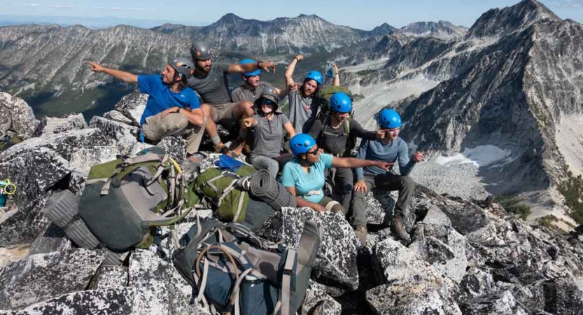 a group of gap year students sit atop a summit and make silly poses. there are mountains in the background